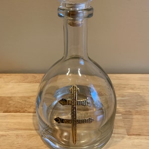 May include: A clear glass bottle with a gold symbol on the front. The bottle has a cork stopper and a clear glass lid.
