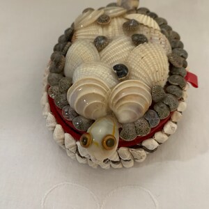 May include: A small, oval-shaped box made of seashells. The box is decorated with a variety of shells, including white, brown, and gray shells. The box has a red lining and a small, white seashell turtle with brown eyes on top.