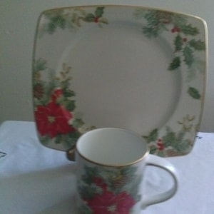 May include: A white ceramic mug and a square plate with a green and red poinsettia floral design. The mug has a gold rim and a handle.