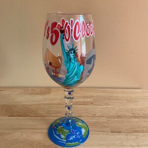 May include: A clear glass wine glass with a blue and green globe design on the base. The glass has a blue stem with blue rhinestones. The glass has a red and white design that says "It's 5 O'Clock Somewhere" with a depiction of the Statue of Liberty.