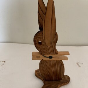May include: Wooden rabbit figurine with long ears and a clothespin attached to its body. The rabbit is brown and has a simple, carved design.