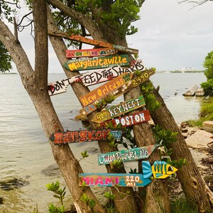 May include: A weathered wooden tree trunk with multiple colourful wooden signs attached to it. The signs point in different directions and have various destinations written on them, including "Bermuda", "Aruba", "Margaritaville", "New Orleans", "Key West", "Boston", "Bangkok", "Outer Banks, NC", and "Miami".