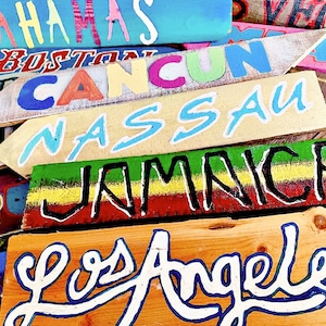 May include: A collection of wooden signs with painted destination names, including "Bahamas", "Boston", "Cancun", "Nassau", "Jamaica", and "Los Angeles". The signs are painted in various bright colours, including blue, green, yellow, red, and white.