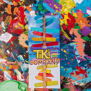 May include: A colourful, abstract background with a white sign that says "The Tiki Company". The sign features a wooden stick with multiple colourful wooden arrows pointing in different directions.