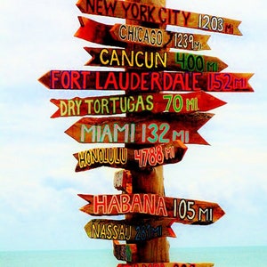 May include: A wooden signpost with multiple directional arrows pointing to different cities and distances. The signpost is weathered and has a rustic look. The arrows are painted in various bright colours and feature the names of cities such as New York City, Chicago, Cancun, Fort Lauderdale, Dry Tortugas, Miami, Honolulu, Habana, Nassau, and Tampa. The distances are listed in miles.