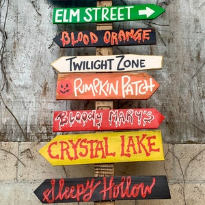 May include: A wooden signpost with multiple directional signs painted in various colours. The signs point to locations like Transylvania, Pet Sematary, Elm Street, Blood Orange, Twilight Zone, Pumpkin Patch, Bloody Mary's, Crystal Lake, Sleepy Hollow, and Haunted Mansion.