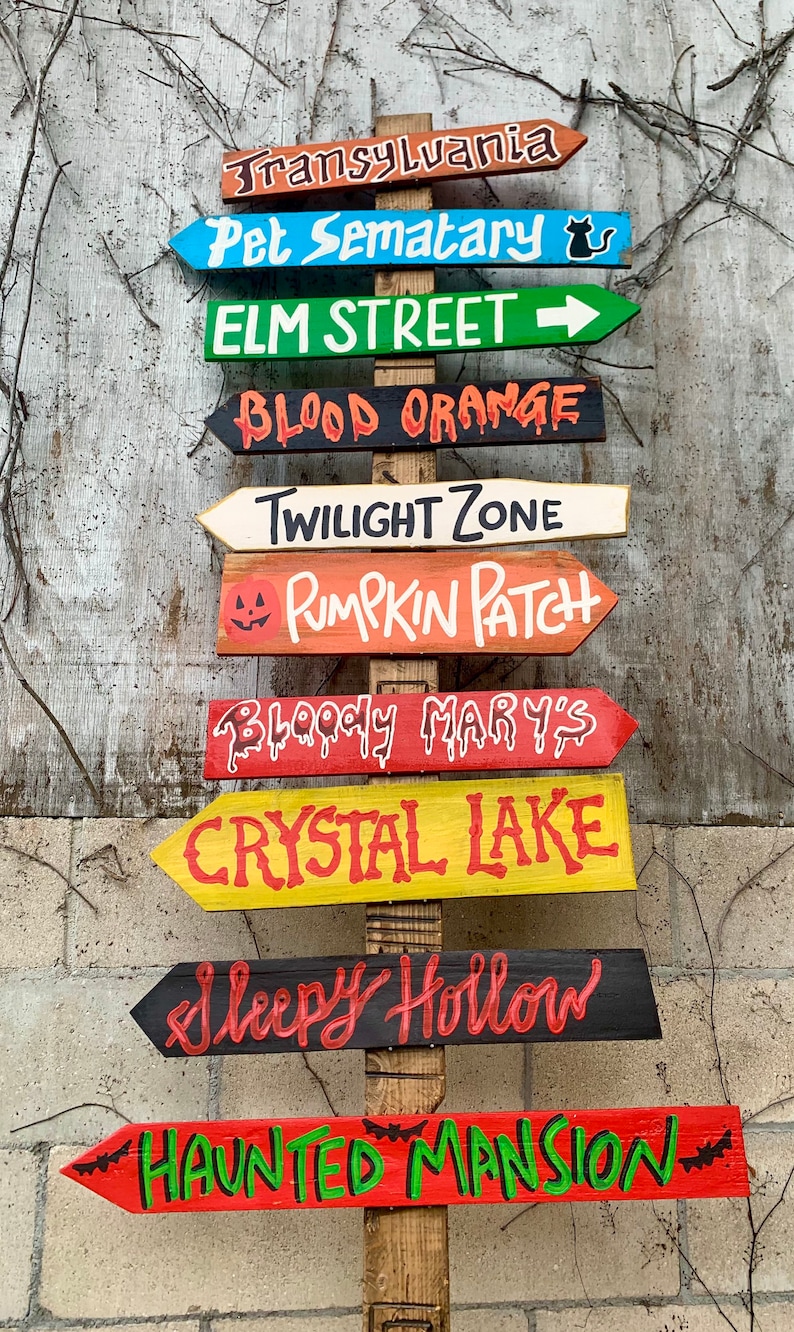 May include: A wooden signpost with multiple directional signs painted in various colours. The signs point to locations like Transylvania, Pet Sematary, Elm Street, Blood Orange, Twilight Zone, Pumpkin Patch, Bloody Mary's, Crystal Lake, Sleepy Hollow, and Haunted Mansion.
