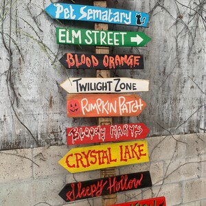 May include: A wooden signpost with multiple directional arrows painted in various colours. Each arrow points to a different spooky location, including Transylvania, Pet Sematary, Elm Street, Blood Orange, Twilight Zone, Pumpkin Patch, Bloody Mary's, Crystal Lake, Sleepy Hollow, and Haunted Mansion.