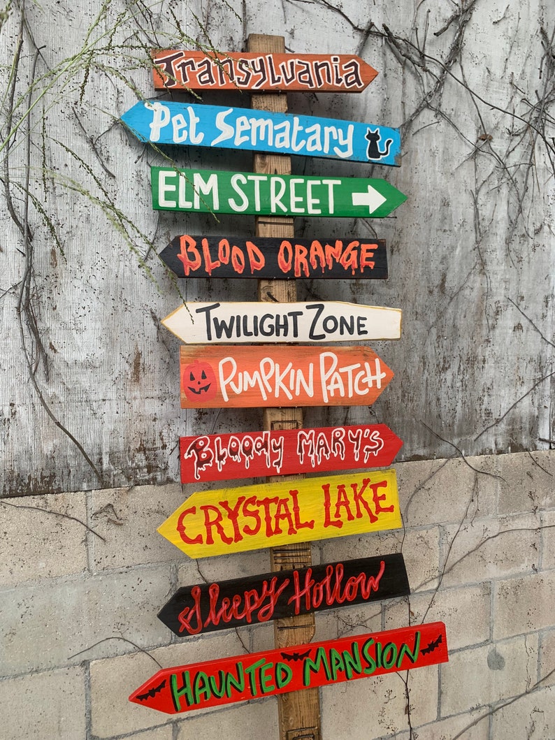 May include: A wooden signpost with multiple directional arrows painted in various colours. Each arrow points to a different spooky location, including Transylvania, Pet Sematary, Elm Street, Blood Orange, Twilight Zone, Pumpkin Patch, Bloody Mary's, Crystal Lake, Sleepy Hollow, and Haunted Mansion.