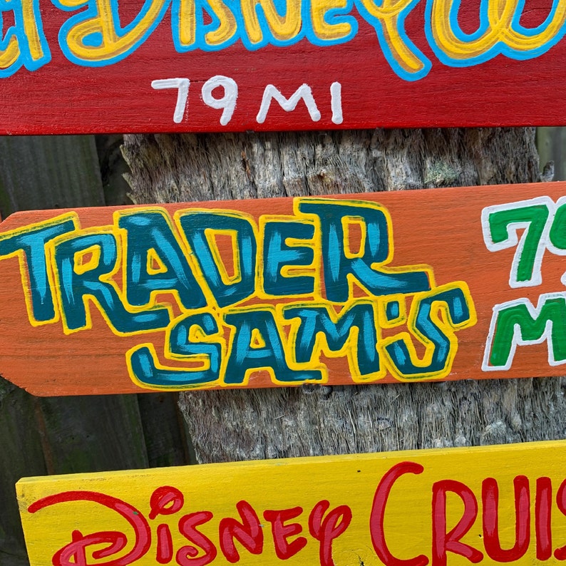 May include: Three hand-painted wooden signs with directional arrows. The top sign is red and says "79 MI". The middle sign is orange and says "TRADER SAM'S". The bottom sign is yellow and says "DISNEY CRUISE".