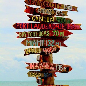 Pu&ograve; includere: Un cartello di legno con pi&ugrave; frecce che puntano in direzioni diverse. Ogni freccia &egrave; dipinta con una destinazione e una distanza diverse, tra cui New York City, Chicago, Cancun, Fort Lauderdale, Dry Tortugas, Miami, Honolulu, L'Avana, Nassau e Tampa.