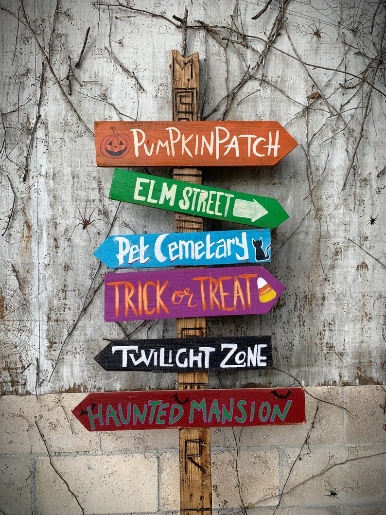 May include: A wooden signpost with six directional arrows pointing to different Halloween destinations. The signs are painted in various colours and feature spooky Halloween-themed text, including "Pumpkin Patch", "Elm Street", "Pet Cemetery", "Trick or Treat", "Twilight Zone", and "Haunted Mansion".