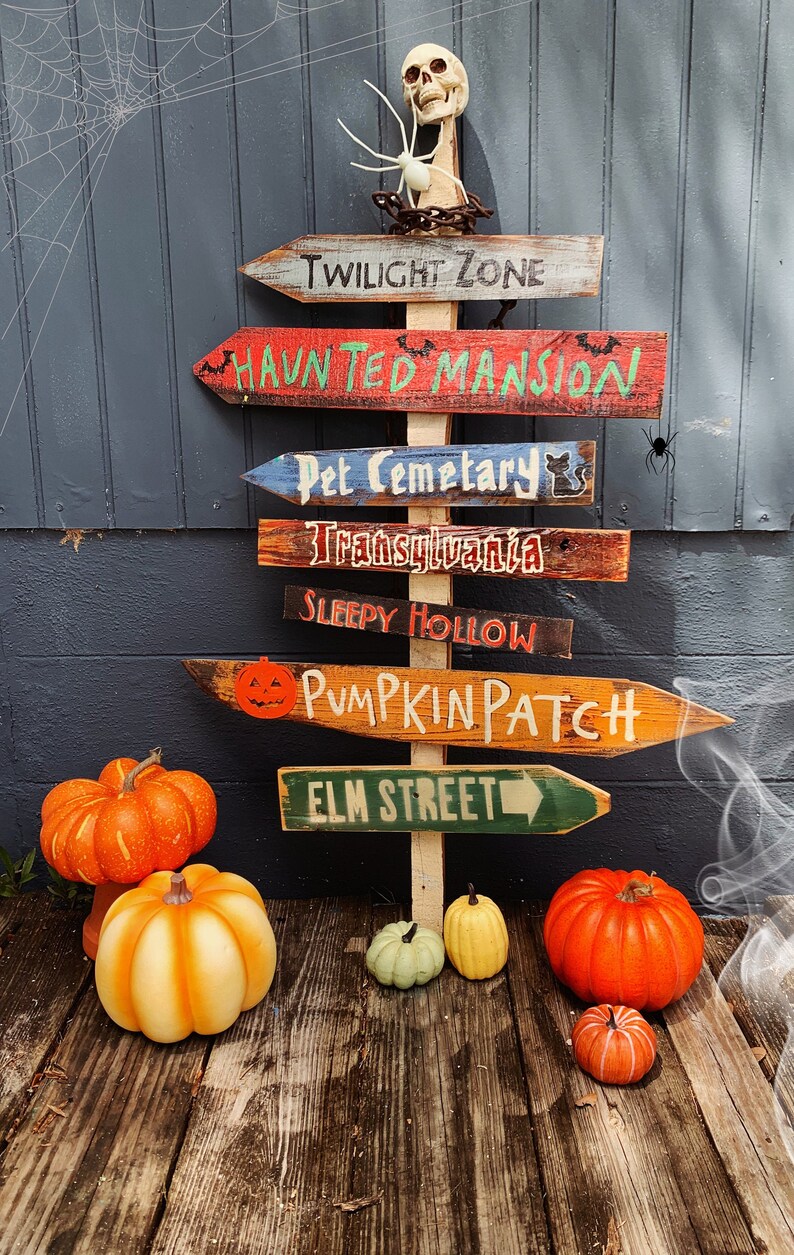 May include: A wooden signpost with multiple directional arrows pointing to various Halloween destinations, including "Twilight Zone", "Haunted Mansion", "Pet Cemetery", "Transylvania", "Sleepy Hollow", "Pumpkin Patch", and "Elm Street". The signpost is decorated with a skull and spider, and there are pumpkins on the ground.