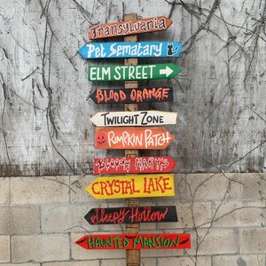 May include: A wooden signpost with multiple directional arrows, each painted in different colours and featuring Halloween-themed destinations like Transylvania, Pet Sematary, Elm Street, Blood Orange, Twilight Zone, Pumpkin Patch, Bloody Mary's, Crystal Lake, Sleepy Hollow, and Haunted Mansion.