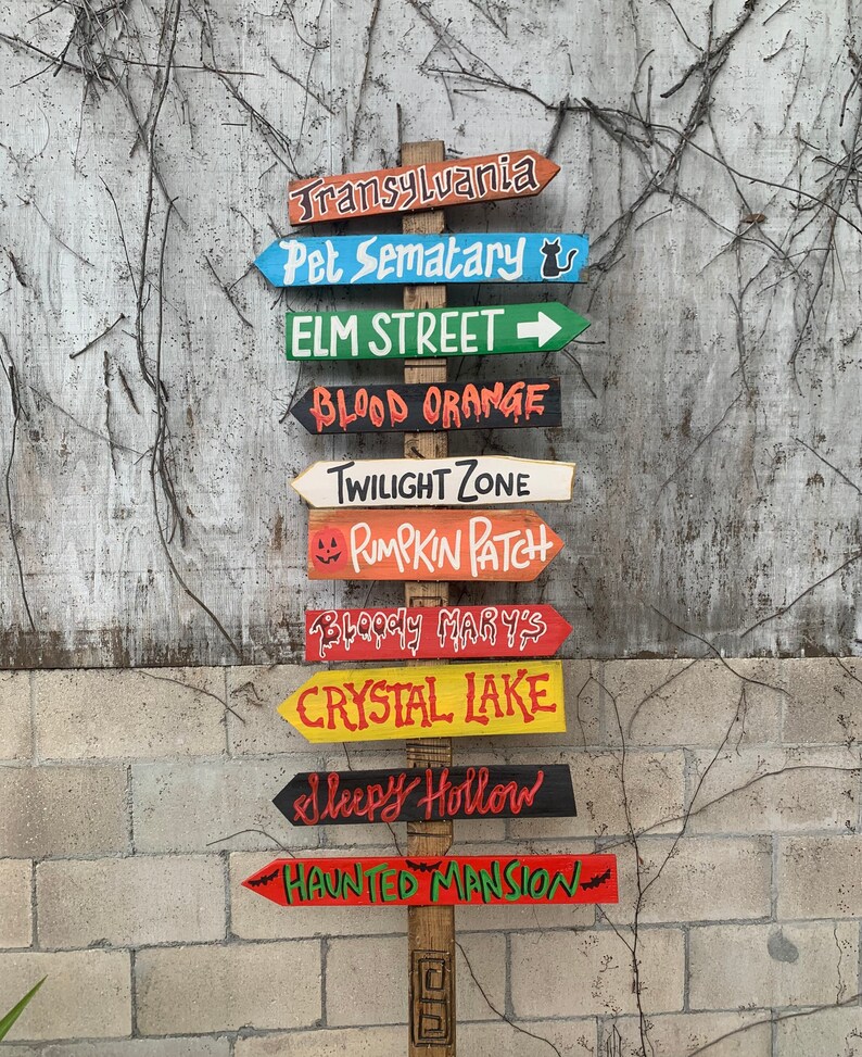 May include: A wooden signpost with multiple directional arrows, each painted in different colours and featuring Halloween-themed destinations like Transylvania, Pet Sematary, Elm Street, Blood Orange, Twilight Zone, Pumpkin Patch, Bloody Mary's, Crystal Lake, Sleepy Hollow, and Haunted Mansion.