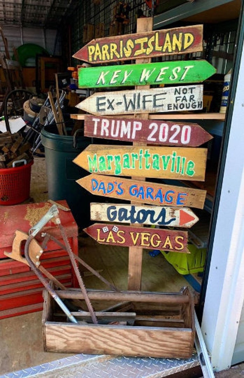 May include: A wooden signpost with multiple arrows pointing in different directions. Each arrow is painted with a different destination, including "Parris Island", "Key West", "Ex-Wife", "Trump 2020", "Margaritaville", "Dad's Garage", "Gators", and "Las Vegas".