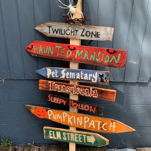 May include: A wooden signpost with multiple directional signs pointing to different Halloween destinations, including "Twilight Zone", "Haunted Mansion", "Pet Sematary", "Transylvania", "Sleepy Hollow", "Pumpkin Patch", and "Elm Street". A skull and spider are at the top of the signpost.