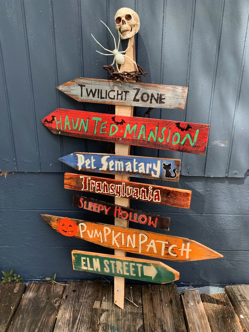 May include: A wooden signpost with multiple directional signs pointing to different Halloween destinations, including "Twilight Zone", "Haunted Mansion", "Pet Sematary", "Transylvania", "Sleepy Hollow", "Pumpkin Patch", and "Elm Street". A skull and spider are at the top of the signpost.