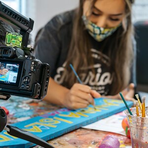 May include: A close-up of a camera filming a person painting a wooden sign with the word "Gloria" on it. The person is wearing a face mask and a grey shirt with the word "Gloria" on it. There are paintbrushes and paint in the background.