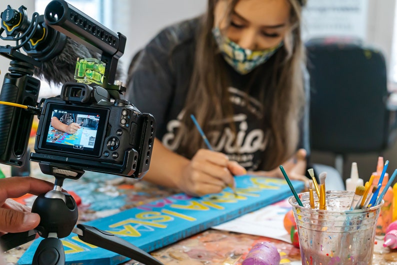 May include: A close-up of a camera filming a person painting a wooden sign with the word "Gloria" on it. The person is wearing a face mask and a grey shirt with the word "Gloria" on it. There are paintbrushes and paint in the background.