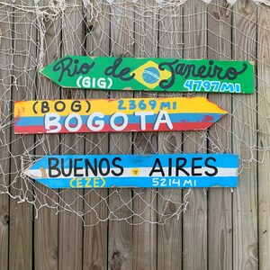 May include: Three wooden signs with painted arrows pointing to the right. The top sign is green and says "Rio de Janeiro (GIG) 4797 MI". The middle sign is yellow and says "(BOG) 2369 MI BOGOTA". The bottom sign is blue and says "BUENOS AIRES (EZE) 5214 MI".
