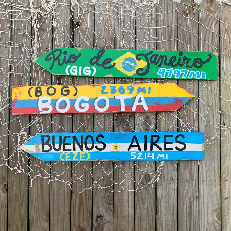 May include: Three wooden signs with painted arrows pointing to the right. The top sign is green and says "Rio de Janeiro (GIG) 4797 MI". The middle sign is yellow and says "(BOG) 2369 MI BOGOTA". The bottom sign is blue and says "BUENOS AIRES (EZE) 5214 MI".