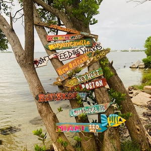 Pu&ograve; includere: Un tronco d'albero di legno consumato con diversi cartelli di legno colorati attaccati. I cartelli puntano in direzioni diverse e hanno varie destinazioni scritte su di essi, tra cui "Bermuda", "Aruba", "Margaritaville", "New Orleans", "Key West", "Boston", "Bangkok", "Outer Banks, NC" e "Miami".