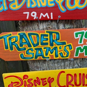 Pu&ograve; includere: Tre cartelli di legno con testo dipinto a mano. Il cartello superiore &egrave; rosso con testo bianco che dice "79 MI". Il cartello centrale &egrave; arancione con testo blu e verde che dice "TRADER SAM'S". Il cartello inferiore &egrave; giallo con testo rosso che dice "DISNEY CRUISE".
