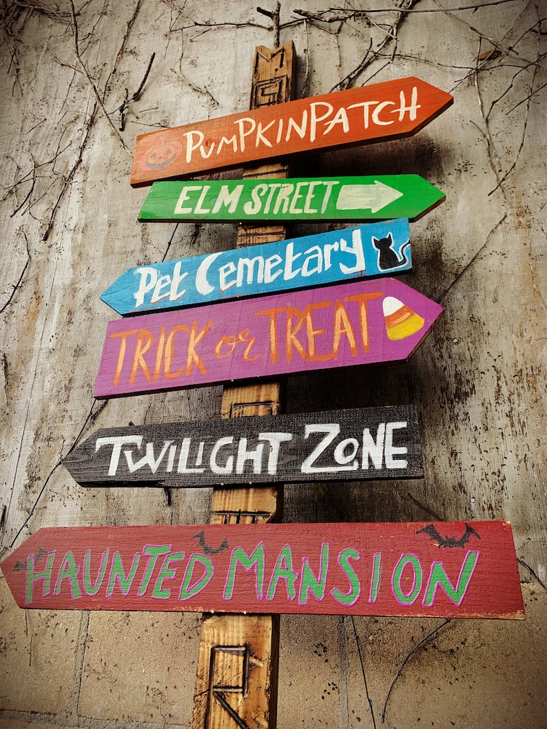 May include: A wooden signpost with six colourful arrows pointing in different directions. The arrows are painted with Halloween-themed words, including "Pumpkin Patch", "Elm Street", "Pet Cemetery", "Trick or Treat", "Twilight Zone", and "Haunted Mansion".