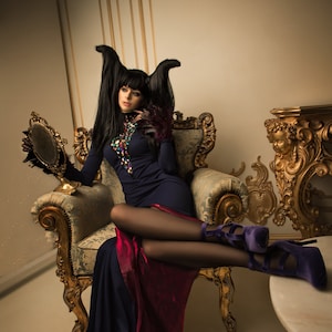May include: A woman with long black hair and horns sits in a gold ornate chair wearing a dark blue dress with a red underskirt and purple heels. She is holding a hand mirror.