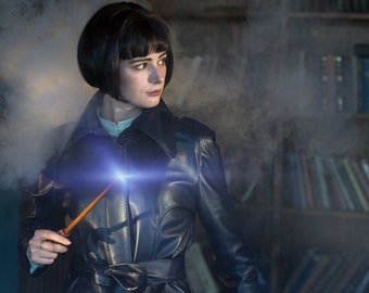 May include: A woman in a black leather trench coat holds a wooden wand in front of a bookshelf. The wand is glowing with a blue light. There is smoke in the background.