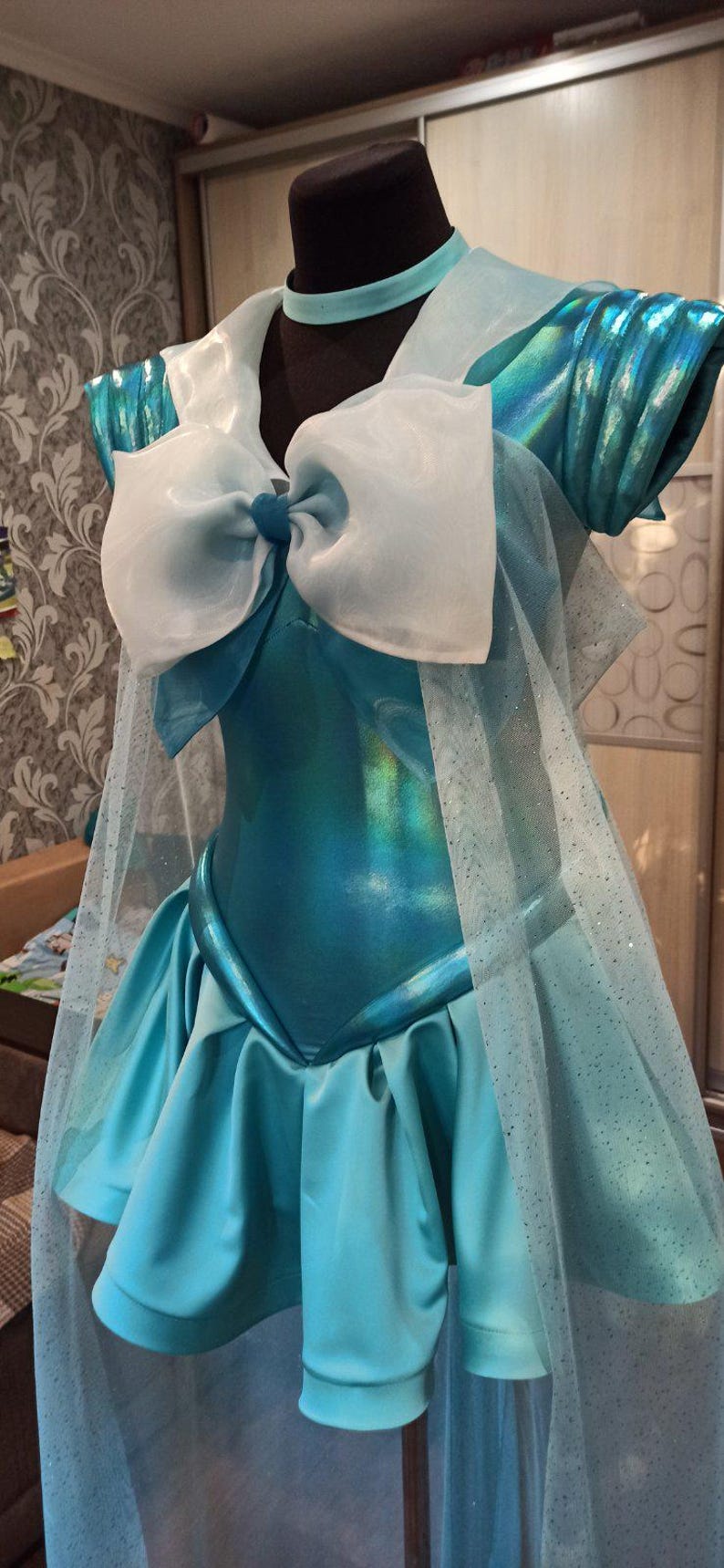 May include: A blue and white cosplay costume with a large white bow and a sheer overlay. The costume is made of a shiny fabric and has a fitted bodice and a full skirt.