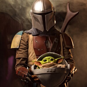 May include: A silver Mandalorian helmet with a brown leather strap and a green alien creature in a gray pod. The Mandalorian is wearing a brown leather vest with a red and brown chest plate. The alien creature is wearing a brown blanket.