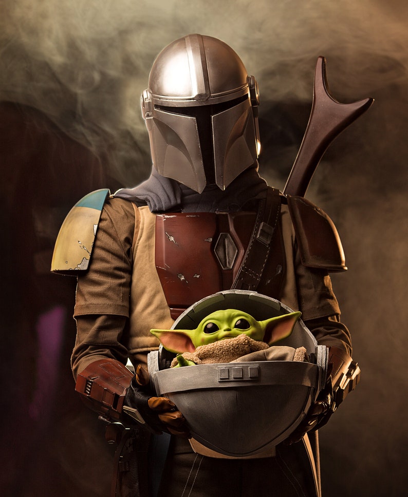 May include: A silver Mandalorian helmet with a brown leather strap and a green alien creature in a gray pod. The Mandalorian is wearing a brown leather vest with a red and brown chest plate. The alien creature is wearing a brown blanket.