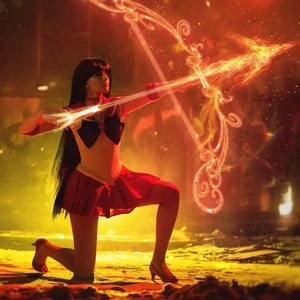 May include: A woman dressed as a character from the anime Sailor Moon, holding a flaming bow and arrow. She is wearing a red skirt, a white top, and a blue bow. The background is a blurry red and yellow.