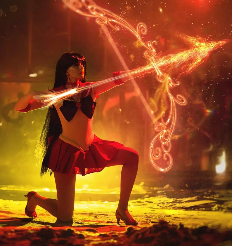 May include: A woman dressed as a character from the anime Sailor Moon, holding a flaming bow and arrow. She is wearing a red skirt, a white top, and a blue bow. The background is a blurry red and yellow.