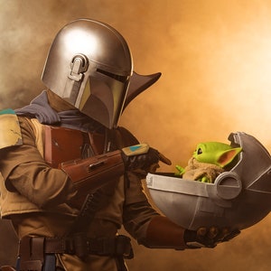 May include: A Mandalorian in a silver and green helmet holds a small, green alien in a floating pod. The alien is wearing a brown robe and has large ears.