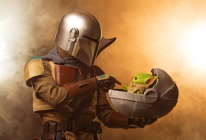 May include: A Mandalorian in a silver and green helmet holds a small, green alien in a floating pod. The alien is wearing a brown robe and has large ears.