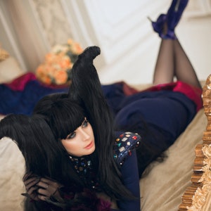 May include: A woman with long black hair styled into horns lies on a bed with a gold headboard. She is wearing a blue and black outfit with a sequined bodice and black gloves. Her legs are crossed and she is wearing high heels.