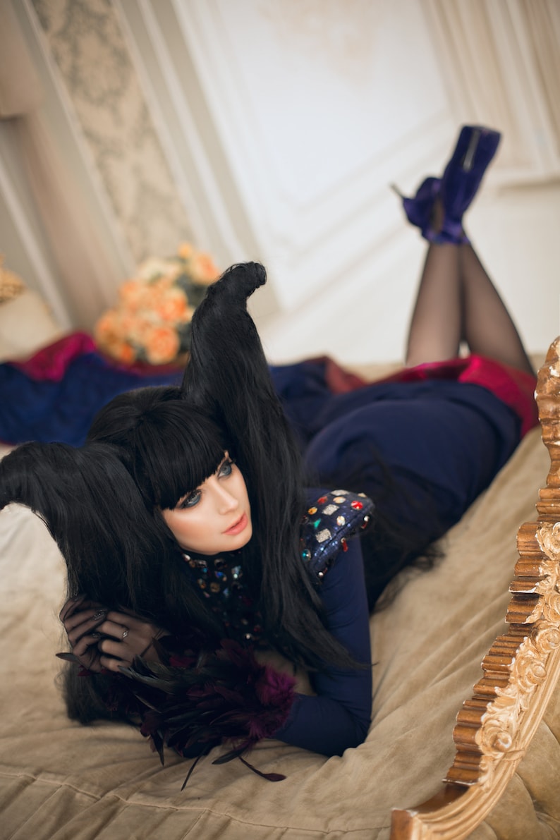 May include: A woman with long black hair styled into horns lies on a bed with a gold headboard. She is wearing a blue and black outfit with a sequined bodice and black gloves. Her legs are crossed and she is wearing high heels.