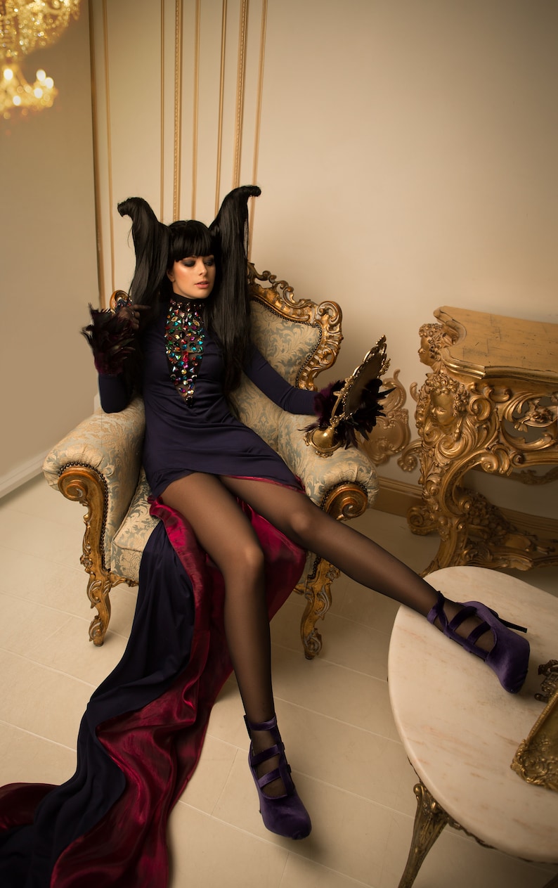 May include: A woman in a dark blue dress with a long train sits in a gold and white armchair. She is wearing black tights and purple heels. The chair is in a room with gold walls and a gold table.