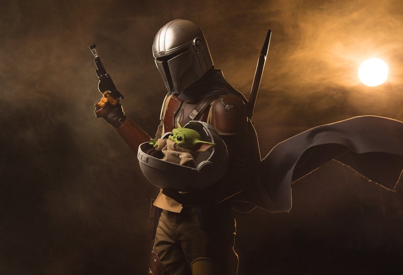 May include: A Mandalorian warrior in a silver helmet and brown armor holds a small green alien in a floating pod. The warrior is holding a blaster pistol in one hand and has a cape flowing behind him. The scene is set against a dark background with a bright orange light in the distance.