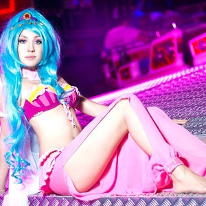 May include: A woman with long blue hair is wearing a pink and white costume with a blue and white top. She is sitting on a pink and silver surface.