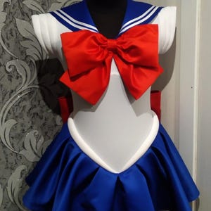 May include: A white and blue sailor-style dress with a red bow. The dress has a white collar with two blue stripes and a white trim around the neckline. The skirt is blue with white trim and has a pleated design.