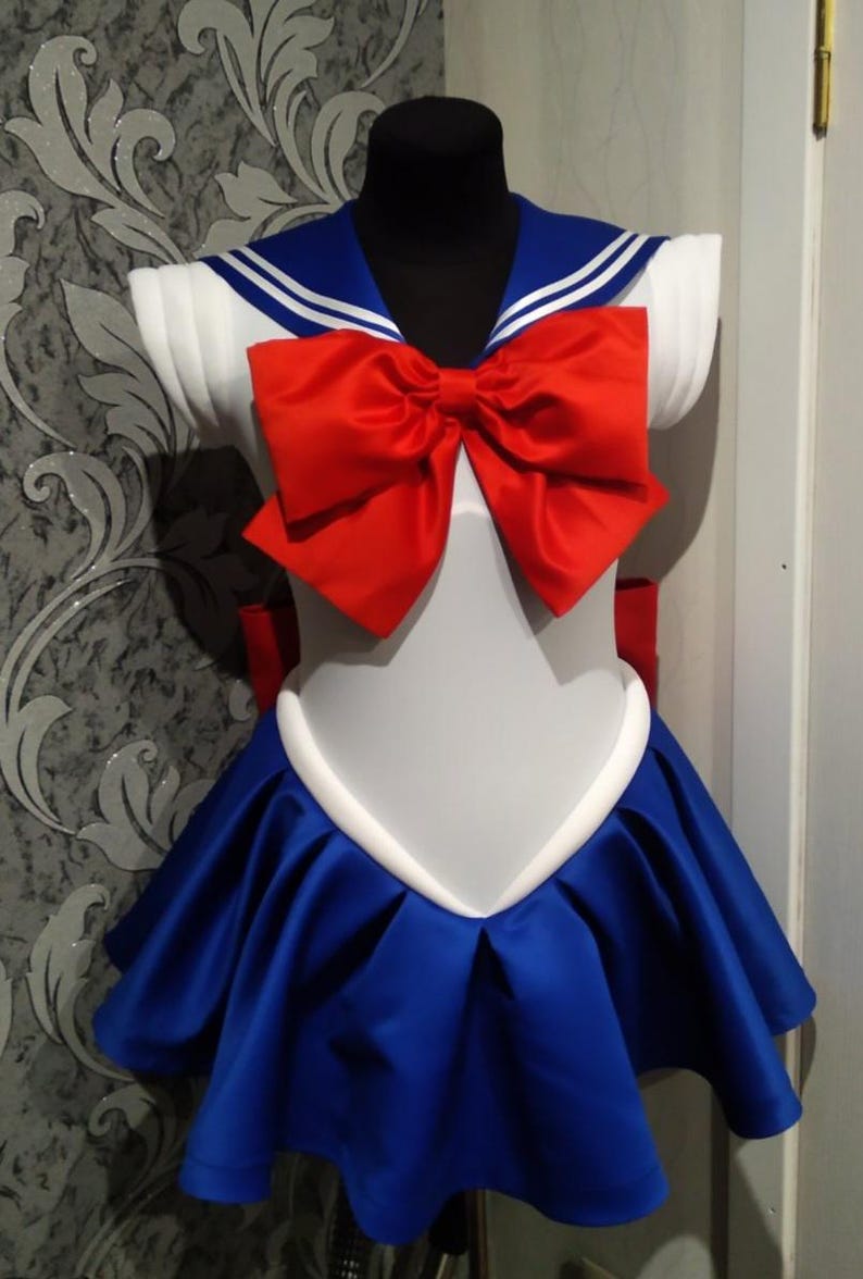 May include: A white and blue sailor-style dress with a red bow. The dress has a white collar with two blue stripes and a white trim around the neckline. The skirt is blue with white trim and has a pleated design.