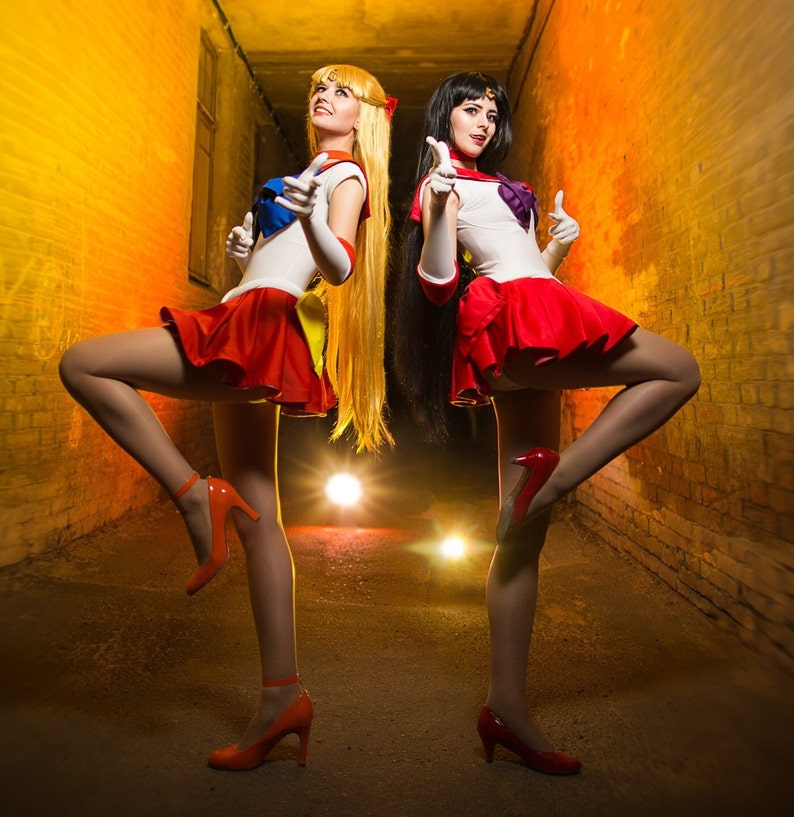 May include: Two women dressed as Sailor Moon characters, one in red and one in blue, pose in a dark alleyway with brick walls. They are both wearing short skirts, white tops, and red high heels.
