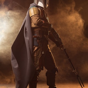 May include: A figure wearing a silver and black helmet and a dark grey cape, holding a long, black weapon with a three-pronged tip. The figure is standing on a brown surface with a smoky background.