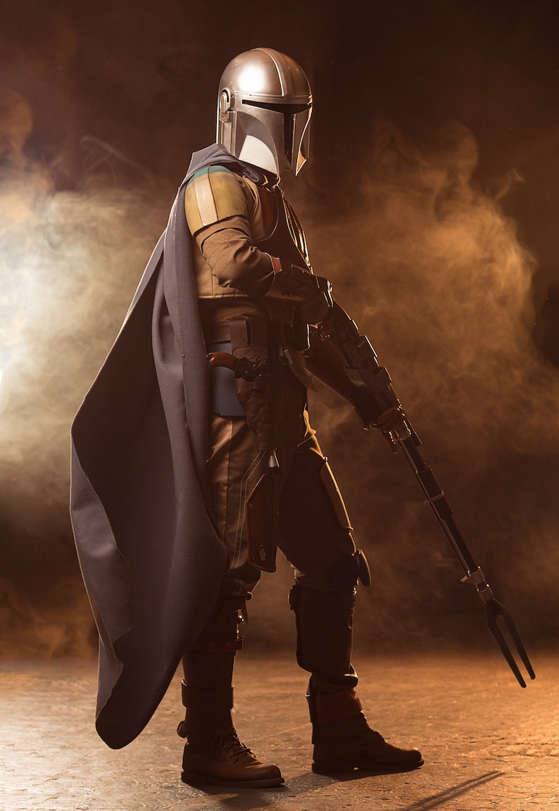May include: A figure wearing a silver and black helmet and a dark grey cape, holding a long, black weapon with a three-pronged tip. The figure is standing on a brown surface with a smoky background.