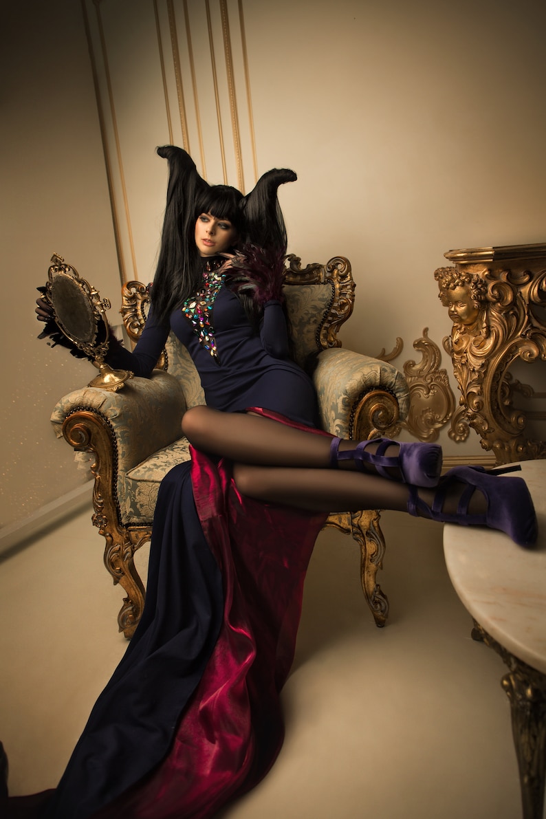 May include: A woman with long black hair and horns sits in a gold ornate chair wearing a dark blue dress with a red underskirt and purple heels. She is holding a hand mirror.