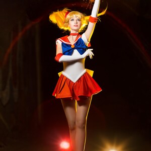 May include: A woman dressed as a Sailor Moon character with a red and orange skirt, white top, and blue bow. She is holding her hands up in the air, and there are glowing hearts around her.